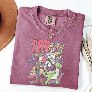 K&ouml;nnte beinhalten: Ein weinrotes T-Shirt mit einem Aufdruck von Woody, Buzz Lightyear und Rex aus dem Film Toy Story. Der Text "TOY STORY" und "SINCE 1995" sind ebenfalls aufgedruckt. Eine goldene Halskette liegt auf dem T-Shirt.