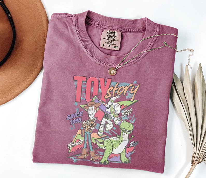 K&ouml;nnte beinhalten: Ein weinrotes T-Shirt mit einem Aufdruck von Woody, Buzz Lightyear und Rex aus dem Film Toy Story. Der Text "TOY STORY" und "SINCE 1995" sind ebenfalls aufgedruckt. Eine goldene Halskette liegt auf dem T-Shirt.