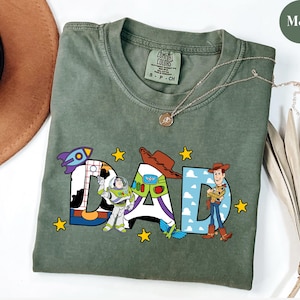 May include: Moss green t-shirt featuring the word "DAD" in colourful cartoon lettering. The letters are decorated with images of a rocket ship, Buzz Lightyear, and Woody from Toy Story. A gold necklace is draped across the shirt.