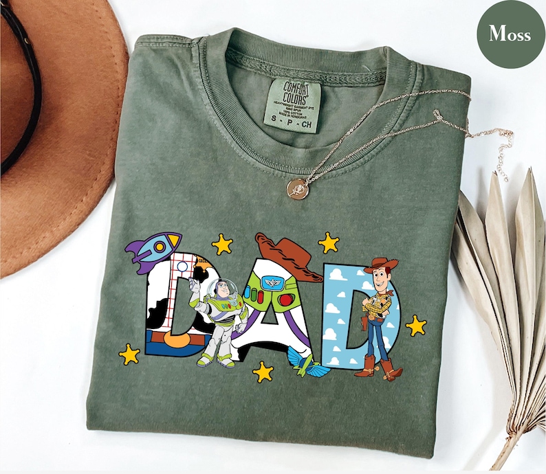 May include: Moss green t-shirt featuring the word "DAD" in colourful cartoon lettering. The letters are decorated with images of a rocket ship, Buzz Lightyear, and Woody from Toy Story. A gold necklace is draped across the shirt.