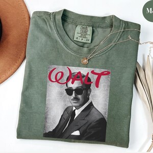 Op de afbeelding: Mosgroen t-shirt met een zwart-wit afbeelding van Walt Disney met een zonnebril, met het woord "WALT" in rode letters. Het shirt heeft een ronde hals en korte mouwen. Een gouden ketting is over het shirt gedrapeerd.