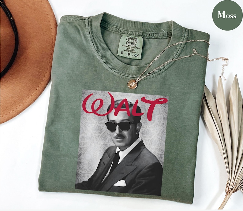 Op de afbeelding: Mosgroen t-shirt met een zwart-wit afbeelding van Walt Disney met een zonnebril, met het woord "WALT" in rode letters. Het shirt heeft een ronde hals en korte mouwen. Een gouden ketting is over het shirt gedrapeerd.