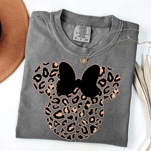 May include: A gray t-shirt featuring a Minnie Mouse design with a black bow and a leopard print pattern. The shirt has a crew neck and short sleeves. A silver necklace with a small pendant is draped across the shirt.