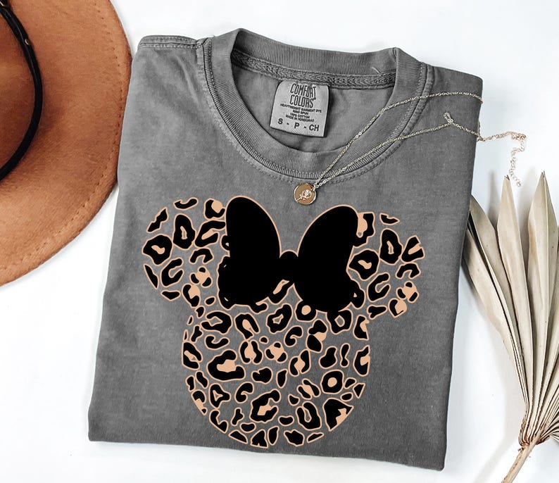 May include: A gray t-shirt featuring a Minnie Mouse design with a black bow and a leopard print pattern. The shirt has a crew neck and short sleeves. A silver necklace with a small pendant is draped across the shirt.