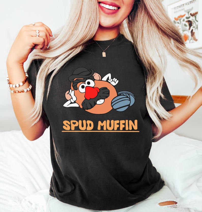 Puede incluir: Camiseta negra con un personaje de dibujos animados de patata con bigote, sombrero y voleibol. El texto "SPUD MUFFIN" est&aacute; impreso en naranja. Un estilo de camisa casual y c&oacute;modo.