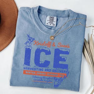 May include: A blue Comfort Colors t-shirt featuring the text "Kristoff & Sven's ICE" in blue and orange, with a graphic of Olaf the snowman and a reindeer. A gold necklace with a pendant is draped across the shirt.