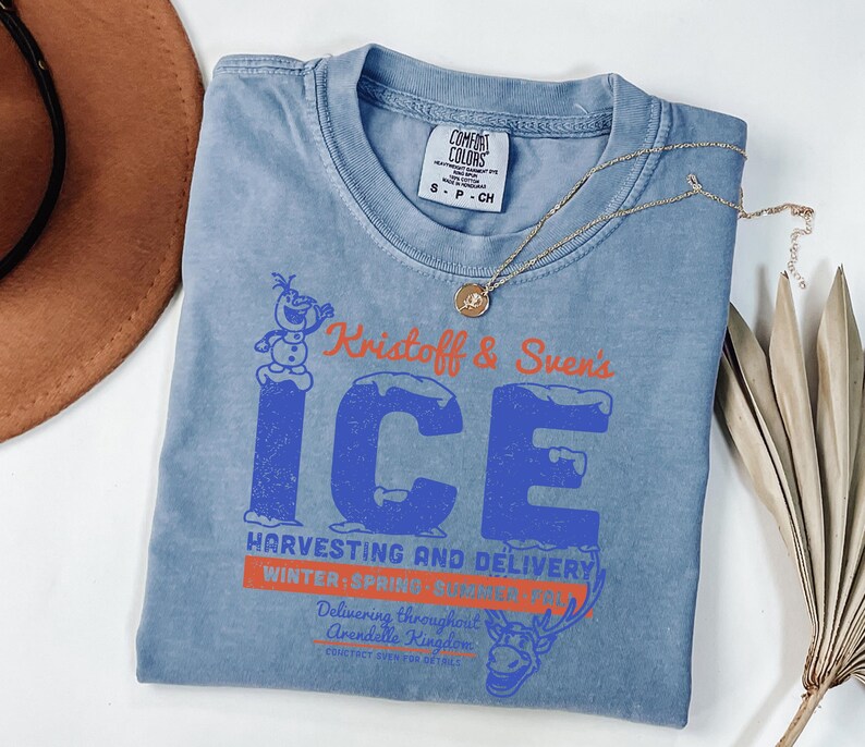May include: A blue Comfort Colors t-shirt featuring the text "Kristoff & Sven's ICE" in blue and orange, with a graphic of Olaf the snowman and a reindeer. A gold necklace with a pendant is draped across the shirt.
