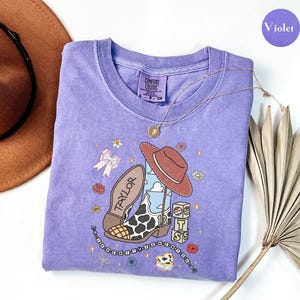 Puede incluir: Una camiseta violeta de manga corta con un estampado gr&aacute;fico de una bota de vaquero con manchas de vaca, un sombrero marr&oacute;n, el nombre TAYLOR, el n&uacute;mero 13 y bloques apilados con las letras T, S y B, decorada con peque&ntilde;as flores y estrellas.