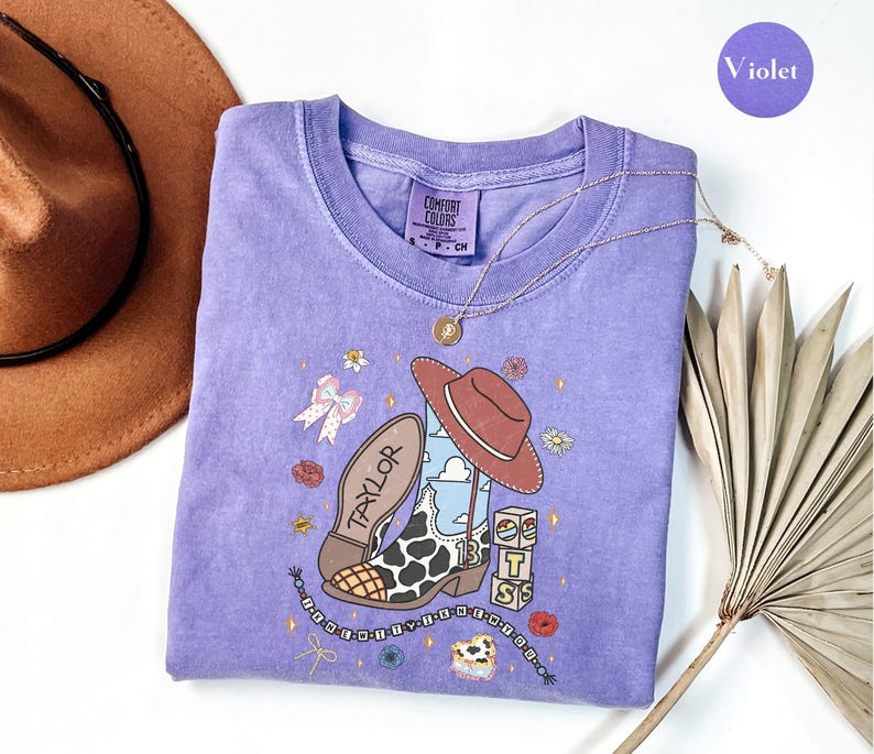 Puede incluir: Una camiseta violeta de manga corta con un estampado gr&aacute;fico de una bota de vaquero con manchas de vaca, un sombrero marr&oacute;n, el nombre TAYLOR, el n&uacute;mero 13 y bloques apilados con las letras T, S y B, decorada con peque&ntilde;as flores y estrellas.