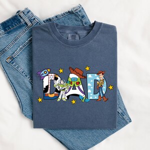 May include: A navy blue t-shirt featuring the word "DAD" in a colourful design with characters from Toy Story. The shirt is folded on top of a pair of blue denim jeans.