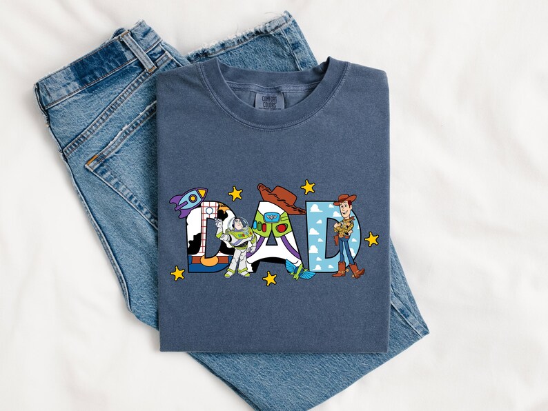 May include: A navy blue t-shirt featuring the word "DAD" in a colourful design with characters from Toy Story. The shirt is folded on top of a pair of blue denim jeans.