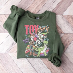 K&ouml;nnte beinhalten: Ein dunkelgr&uuml;nes Sweatshirt mit einem Grafikdesign aus dem Film "Toy Story". Das Design zeigt Woody, Buzz Lightyear, Rex und die Worte "Toy Story" und "Since 1995". Das Sweatshirt hat gerollte &Auml;rmel und einen Rundhalsausschnitt.