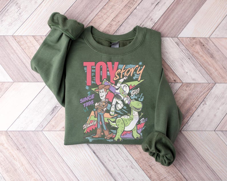 K&ouml;nnte beinhalten: Ein dunkelgr&uuml;nes Sweatshirt mit einem Grafikdesign aus dem Film "Toy Story". Das Design zeigt Woody, Buzz Lightyear, Rex und die Worte "Toy Story" und "Since 1995". Das Sweatshirt hat gerollte &Auml;rmel und einen Rundhalsausschnitt.