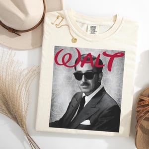 Op de afbeelding: Cr&egrave;mekleurig T-shirt met een zwart-wit afbeelding van een man met een zonnebril en het woord "WALT" in rode letters. Het shirt is gestyled met een hoed, sandalen en een gouden ketting.