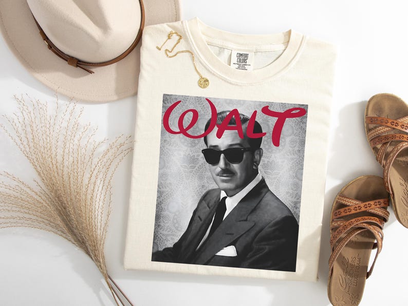 Op de afbeelding: Cr&egrave;mekleurig T-shirt met een zwart-wit afbeelding van een man met een zonnebril en het woord "WALT" in rode letters. Het shirt is gestyled met een hoed, sandalen en een gouden ketting.