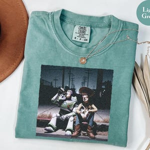 May include: A light green short-sleeve t-shirt featuring a graphic print of Buzz Lightyear and Woody sitting on a curb at night. The shirt has a crew neckline and a Comfort Colors brand label on the inside collar. A thin gold chain necklace rests on the fabric.