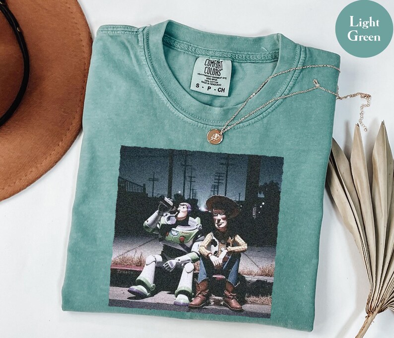 May include: A light green short-sleeve t-shirt featuring a graphic print of Buzz Lightyear and Woody sitting on a curb at night. The shirt has a crew neckline and a Comfort Colors brand label on the inside collar. A thin gold chain necklace rests on the fabric.
