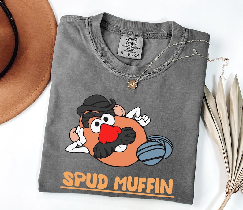 Puede incluir: Camiseta gris con una ilustraci&oacute;n de dibujos animados de un personaje de patata con sombrero y bigote, con el texto "SPUD MUFFIN" en naranja. La camiseta tiene cuello redondo y una etiqueta Comfort Colors.