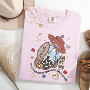 Puede incluir: Una camiseta rosa claro con un estampado gr&aacute;fico de una bota de vaquero con patr&oacute;n de manchas de vaca, un sombrero de vaquero marr&oacute;n, el nombre Taylor, el n&uacute;mero 13 y bloques apilados con las letras T, S y un icono de sol.