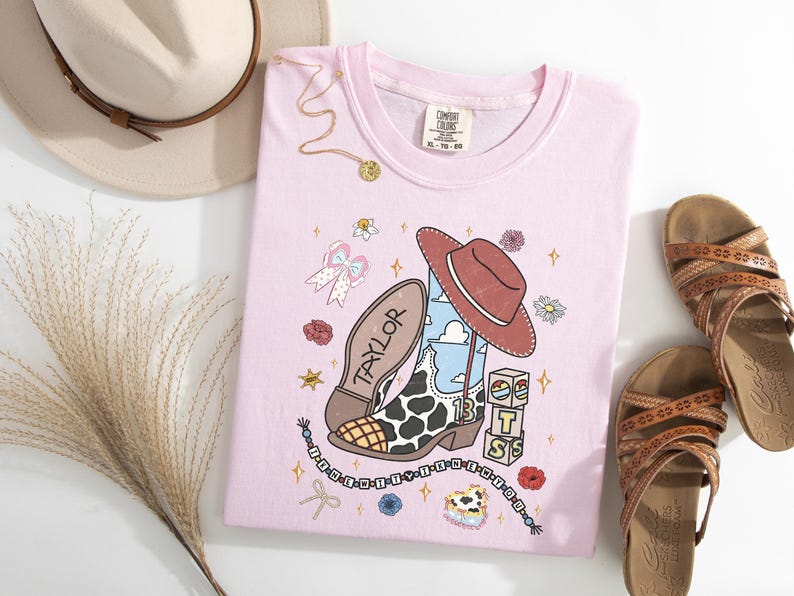 Puede incluir: Una camiseta rosa claro con un estampado gr&aacute;fico de una bota de vaquero con patr&oacute;n de manchas de vaca, un sombrero de vaquero marr&oacute;n, el nombre Taylor, el n&uacute;mero 13 y bloques apilados con las letras T, S y un icono de sol.