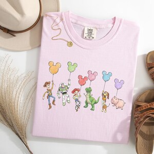 May include: A light pink t-shirt featuring a graphic print of six animated characters holding colourful Mickey Mouse-shaped balloons. The shirt has a Comfort Colors brand label at the neckline. The design includes a cowboy, space ranger, cowgirl, dinosaur, dachshund, and pig character.