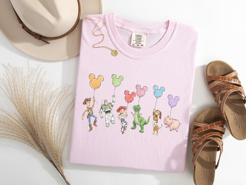 May include: A light pink t-shirt featuring a graphic print of six animated characters holding colourful Mickey Mouse-shaped balloons. The shirt has a Comfort Colors brand label at the neckline. The design includes a cowboy, space ranger, cowgirl, dinosaur, dachshund, and pig character.