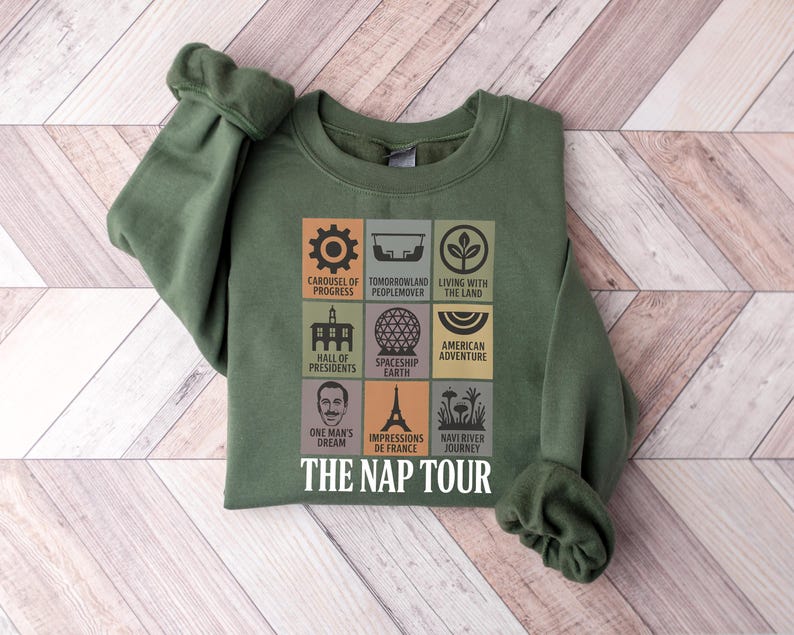 May include: Green sweatshirt with a graphic featuring icons and text from various attractions. The text includes "Carousel of Progress," "Tomorrowland Peoplemover," "Living with the Land," "Hall of Presidents," "Spaceship Earth," "American Adventure," "One Man's Dream," "Impressions de France," "Navi River Journey," and "The Nap Tour."