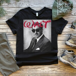 Op de afbeelding: Zwart t-shirt met een grijswaardenafbeelding van een man met een zonnebril en een pak, met de naam "WALT" in rode letters. Het shirt wordt getoond met olijfgroene broek, wandelschoenen, een groene bandana en een kompas.