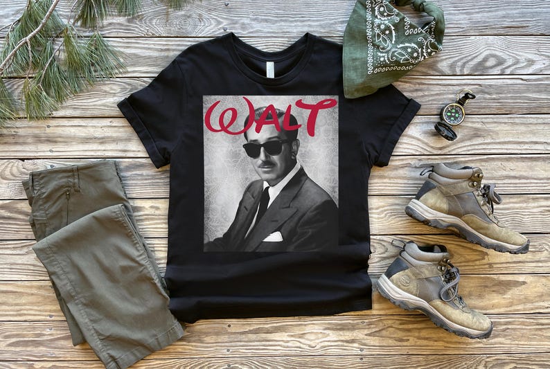 Op de afbeelding: Zwart t-shirt met een grijswaardenafbeelding van een man met een zonnebril en een pak, met de naam "WALT" in rode letters. Het shirt wordt getoond met olijfgroene broek, wandelschoenen, een groene bandana en een kompas.