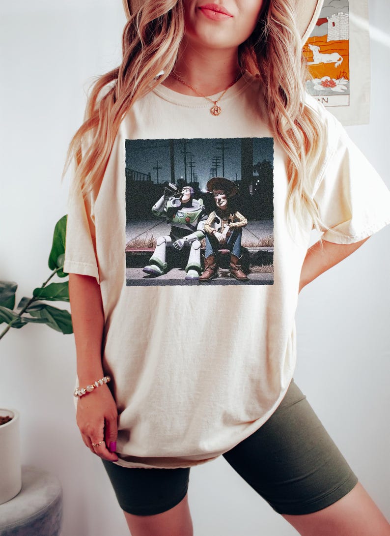May include: A cream-colored short-sleeve t-shirt featuring a graphic print of the characters Buzz Lightyear and Woody sitting on a curb at night. The graphic shows Buzz Lightyear drinking from a bottle while Woody sits beside him.