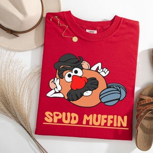 Puede incluir: Una camiseta roja con un personaje de dibujos animados de patata con sombrero negro y bigote, y una nariz roja. El texto "SPUD MUFFIN" est&aacute; impreso en naranja. Un sombrero beige, sandalias y joyas tambi&eacute;n est&aacute;n en la imagen.