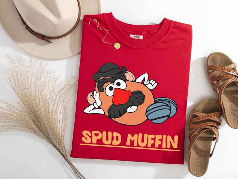 Puede incluir: Una camiseta roja con un personaje de dibujos animados de patata con sombrero negro y bigote, y una nariz roja. El texto "SPUD MUFFIN" est&aacute; impreso en naranja. Un sombrero beige, sandalias y joyas tambi&eacute;n est&aacute;n en la imagen.