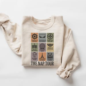 May include: Cream-colored sweatshirt featuring a colorful graphic of themed attractions. The design includes icons and text such as "Carousel of Progress," "Spaceship Earth," and "The Nap Tour." A comfortable, casual top.