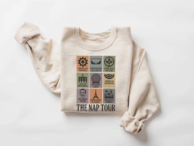 May include: Cream-colored sweatshirt featuring a colorful graphic of themed attractions. The design includes icons and text such as "Carousel of Progress," "Spaceship Earth," and "The Nap Tour." A comfortable, casual top.