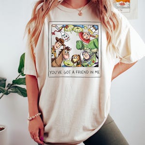 Puede incluir: Camiseta color crema con una colorida ilustraci&oacute;n de personajes de la pel&iacute;cula animada "Toy Story". La imagen incluye a Woody, Buzz Lightyear y otros personajes. El texto "YOU'VE GOT A FRIEND IN ME" est&aacute; impreso debajo.