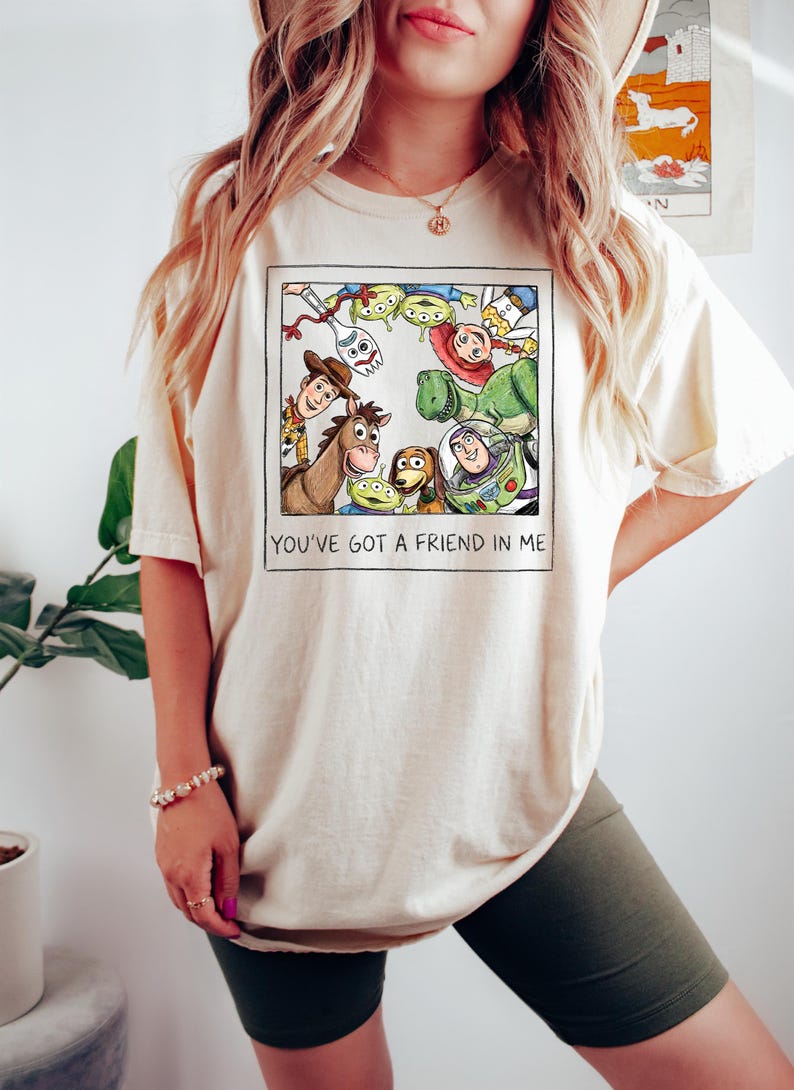 Puede incluir: Camiseta color crema con una colorida ilustraci&oacute;n de personajes de la pel&iacute;cula animada "Toy Story". La imagen incluye a Woody, Buzz Lightyear y otros personajes. El texto "YOU'VE GOT A FRIEND IN ME" est&aacute; impreso debajo.