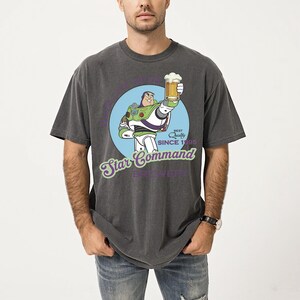 May include: Dark gray t-shirt featuring a cartoon Buzz Lightyear holding a beer, with the text "Star Command Brewery" and "Best Quality Since 1995". The design is set in a blue circle. The shirt is a relaxed fit.