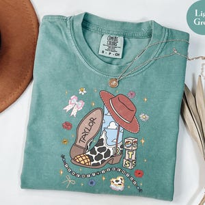 Puede incluir: Una camiseta de manga corta de color verde claro con un estampado gr&aacute;fico de una bota de vaquero con punta de estampado de vaca, un sombrero marr&oacute;n, bloques con las letras T, S y un arco&iacute;ris, un lazo y una cadena de cuentas con el texto I KNEW YOU WERE TROUBLE.