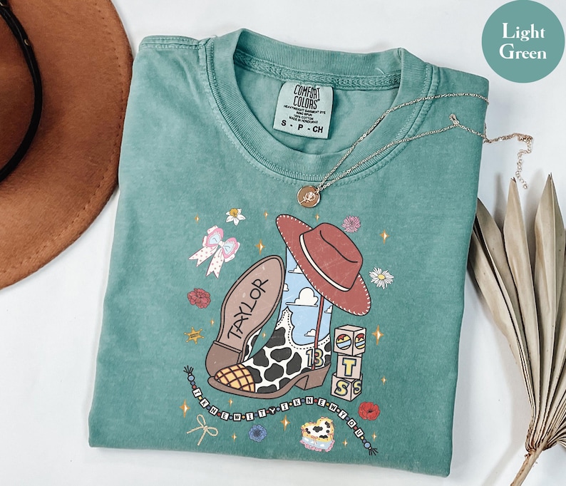 Puede incluir: Una camiseta de manga corta de color verde claro con un estampado gr&aacute;fico de una bota de vaquero con punta de estampado de vaca, un sombrero marr&oacute;n, bloques con las letras T, S y un arco&iacute;ris, un lazo y una cadena de cuentas con el texto I KNEW YOU WERE TROUBLE.
