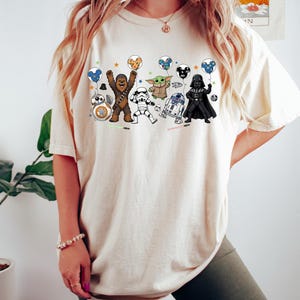 May include: A cream-colored t-shirt featuring a graphic print of various Star Wars characters including Chewbacca, a Stormtrooper, Grogu, R2-D2, Darth Vader, and BB-8, each holding a colorful balloon with a Mickey Mouse ear silhouette design.