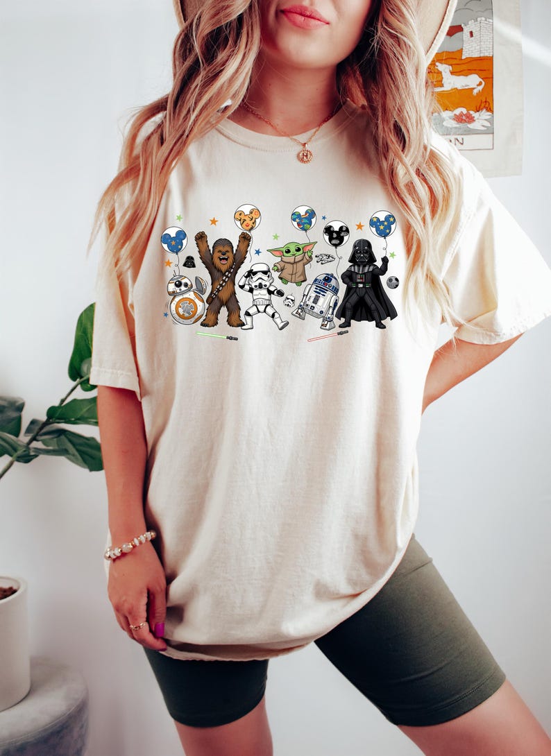 May include: A cream-colored t-shirt featuring a graphic print of various Star Wars characters including Chewbacca, a Stormtrooper, Grogu, R2-D2, Darth Vader, and BB-8, each holding a colorful balloon with a Mickey Mouse ear silhouette design.