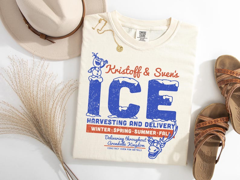May include: Cream-colored t-shirt with a graphic featuring the text "Kristoff & Sven's ICE" in blue and red. The design includes a snowman and a reindeer. A hat, sandals, and jewelry are also in the image.