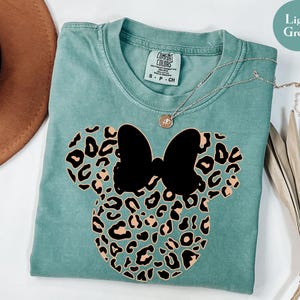 May include: Light green t-shirt featuring a black bow and leopard print mouse ears design. The shirt has a crew neck and short sleeves. A silver necklace with a small pendant is draped across the shirt. The shirt is made of a soft material.