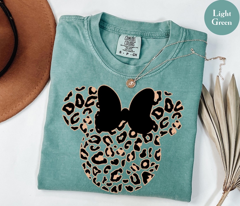 May include: Light green t-shirt featuring a black bow and leopard print mouse ears design. The shirt has a crew neck and short sleeves. A silver necklace with a small pendant is draped across the shirt. The shirt is made of a soft material.