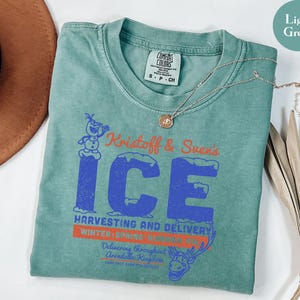 May include: Light green t-shirt featuring a graphic design with the text "Kristoff & Sven's ICE" in blue and orange. The design includes an image of Olaf the snowman and Sven the reindeer. A gold necklace is draped across the shirt.