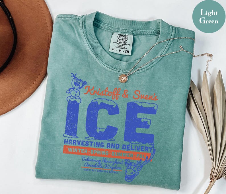 May include: Light green t-shirt featuring a graphic design with the text "Kristoff & Sven's ICE" in blue and orange. The design includes an image of Olaf the snowman and Sven the reindeer. A gold necklace is draped across the shirt.