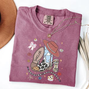 Puede incluir: Una camiseta de manga corta color malva con un estampado gr&aacute;fico de una bota de vaquero con manchas de vaca, un sombrero marr&oacute;n y bloques con el texto "TS". La bota tiene "TAYLOR" en la suela y el n&uacute;mero "13" en el lateral. El dise&ntilde;o incluye flores peque&ntilde;as, estrellas y una cadena de cuentas.