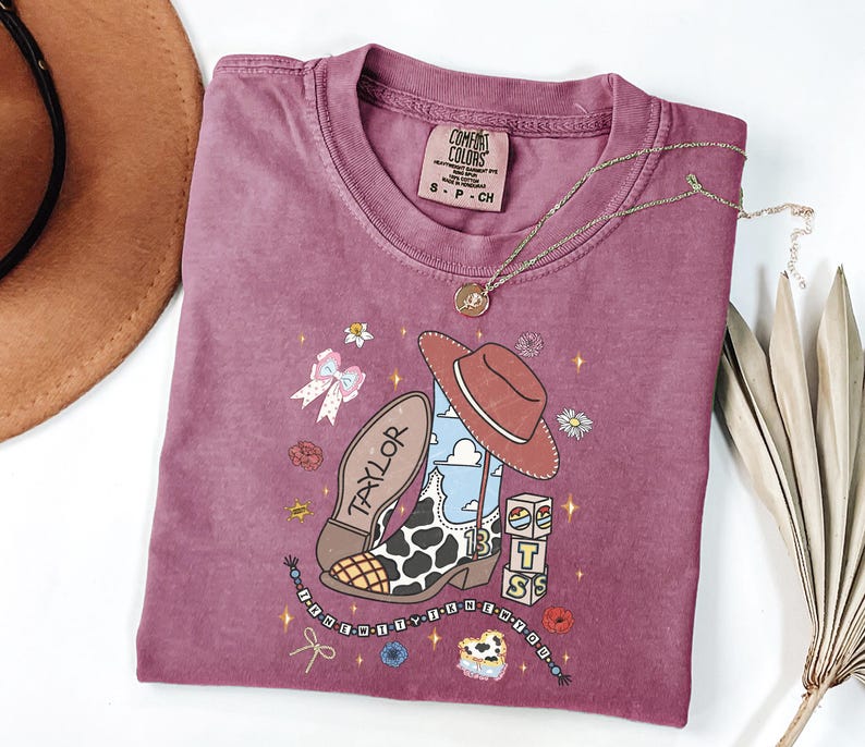 Puede incluir: Una camiseta de manga corta color malva con un estampado gr&aacute;fico de una bota de vaquero con manchas de vaca, un sombrero marr&oacute;n y bloques con el texto "TS". La bota tiene "TAYLOR" en la suela y el n&uacute;mero "13" en el lateral. El dise&ntilde;o incluye flores peque&ntilde;as, estrellas y una cadena de cuentas.