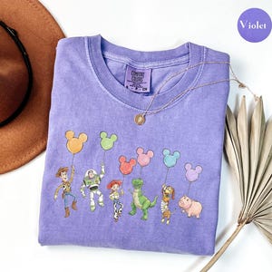 May include: A violet short-sleeved t-shirt featuring a graphic print of six animated characters from Toy Story, each holding a Mickey Mouse-shaped balloon in various colours. The shirt has a Comfort Colors brand label at the inner neckline.