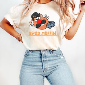Puede incluir: Camiseta color crema con un personaje de dibujos animados de patata con sombrero negro, bigote y nariz roja. El texto "SPUD MUFFIN" est&aacute; impreso en naranja. La persona lleva vaqueros azul claro.